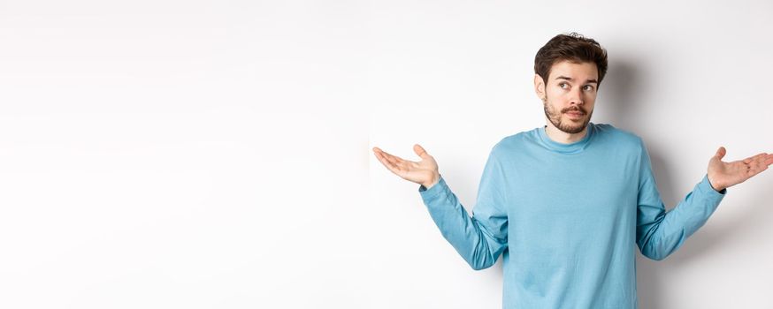 Indecisive young man with beard, shrugging shoulders and know nothing, looking away confused, standing in blue shirt over white background.