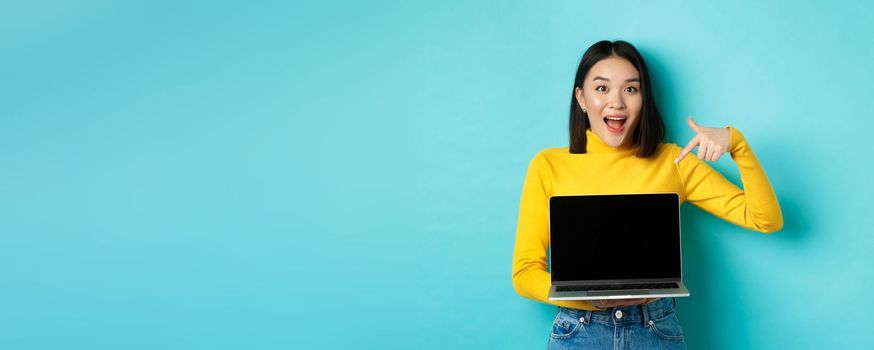 E-commerce and online shopping concept. Excited asian woman making presentation, pointing finger and showing blank laptop screen, standing over blue background.