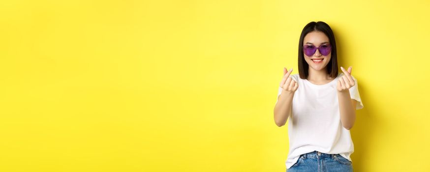 Fashion and lifestyle concept. Attractive asian woman in heart-shape sunglasses, showing finger hearts and smiling happy at camera, standing over yellow background.