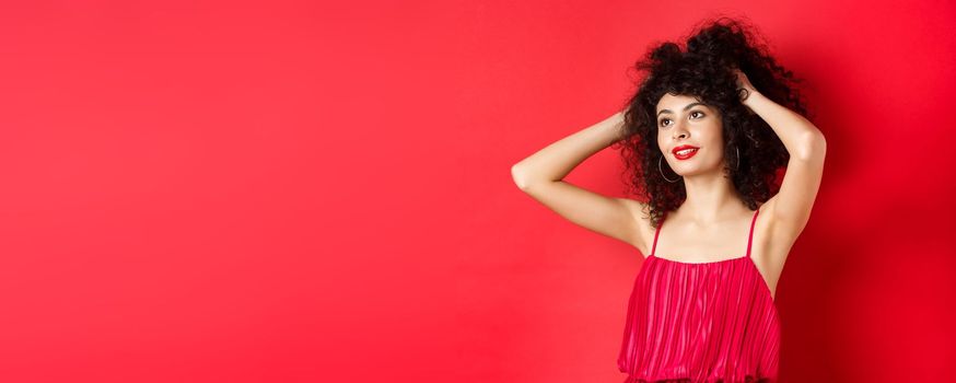 Carefree female model playing with hair, looking dreamy and relaxed at logo, smiling sensual, standing on red background.