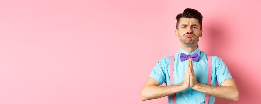 Silly guy making begging face, pleading and asking for help, standing with hands pressed together on pink background. Copy space
