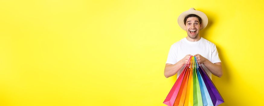 Image of happy man shopping on vacation, holding paper bags and smiling, standing against yellow background.