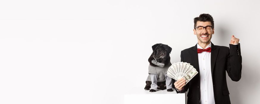 Happy young man and cute black dog standing in party costumes, pug owner holding money dollars and rejoicing, winning prize, white background.