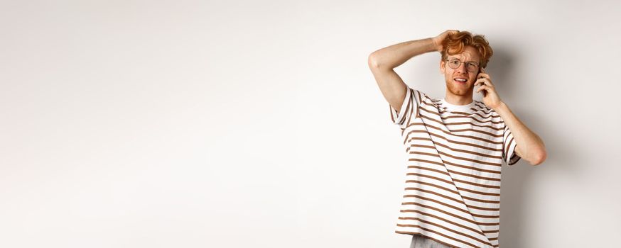 Puzzled redhead guy talking on phone, scratching head and looking confused or indecisive, standing over white background.