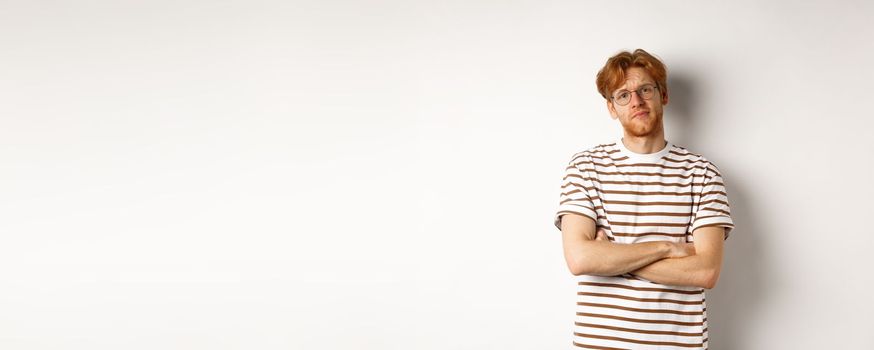 Teenage redhead guy in glasses cross arms on chest, looking skeptical and unamused at camera, standing over white background.