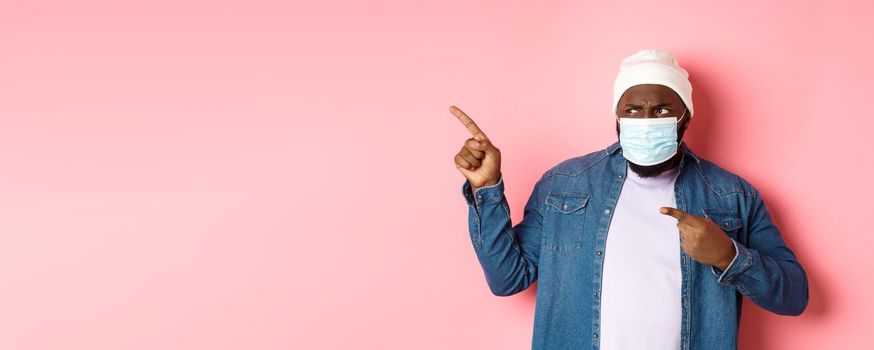 Coronavirus, lifestyle and global pandemic concept. Confused and disappointed african-american guy in face mask pointing fingers left, staring upset with camera, pink background.