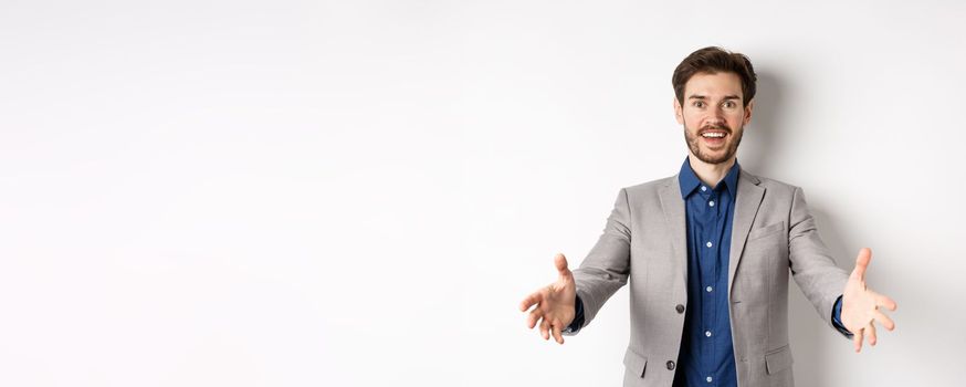 Happy handsome man in suit stretch out hands to greet business partners, welcome guests and smiling excited, standing on white background.