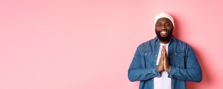 Happy smiling Black man saying thank you, holding hands in pray or namaste gesture, standing grateful against pink background.