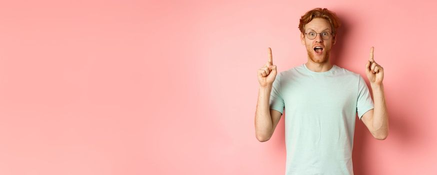 Portrait of surprised guy checking out advertisement, gasping amazed and pointing fingers up, showing promo, standing over pink background.