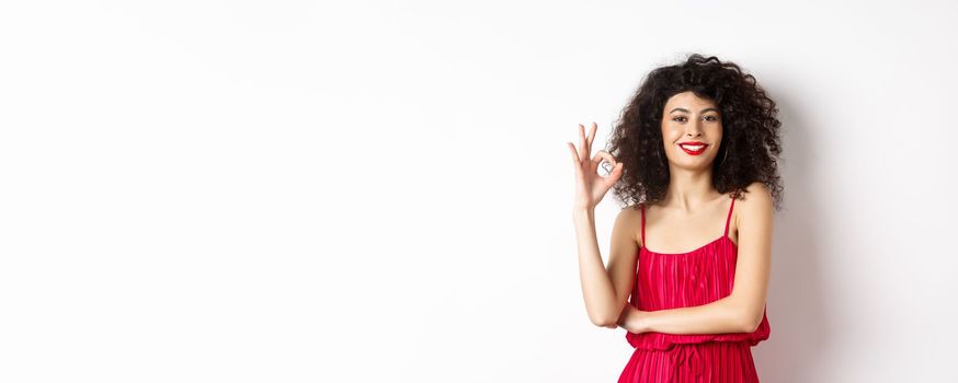 Elegant woman in romantic red dress and makeup, showing okay sign and smiling, recommending good thing, approve and agree, standing satisfied on white background.