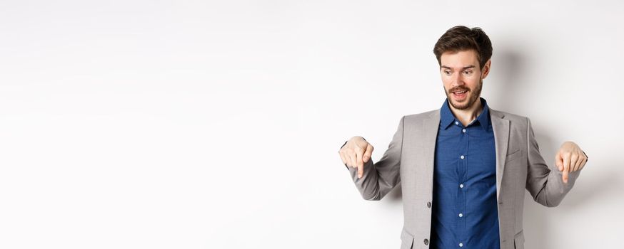 Excited guy in suit smiling and pointing fingers down, looking at special deal with amused face, standing on white background.