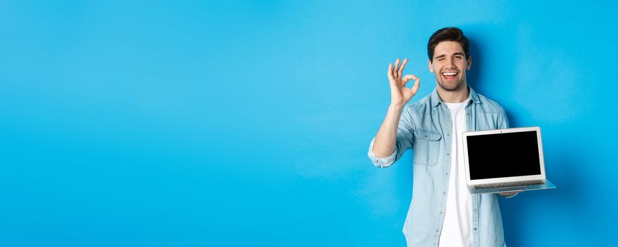 Young man showing laptop screen and okay sign, approve or like promo in internet, smiling satisfied, standing over blue background.