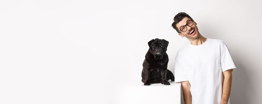 Image of handsome young man standing near cute black pug and smiling. Dog owner spending time with his pet, staring at camera happy, white background.