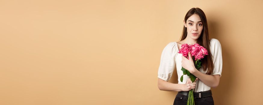 Valentines day. Surprised tender girl thanking for flowers and smiling, holding red roses and smiling grateful, standing on beige background.