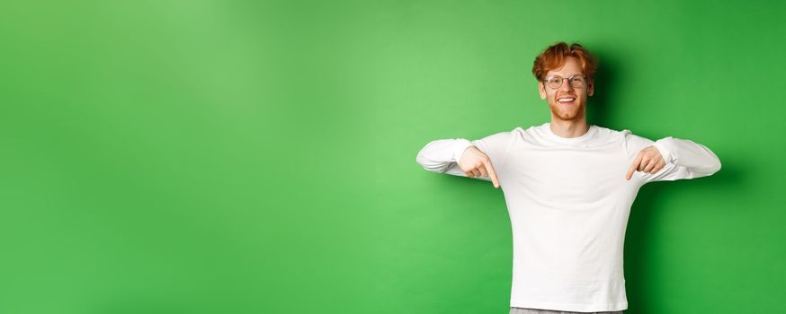 Handsome male model with red hair, wearing glasses and long-sleeve, pointing fingers down and smiling cheerful, showing promo, green background.