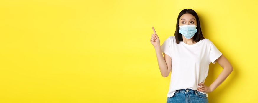 Covid, health care and pandemic concept. Asian woman in medical mask and white t-shirt pointing finger at upper left corner logo, showing promotion, yellow background.