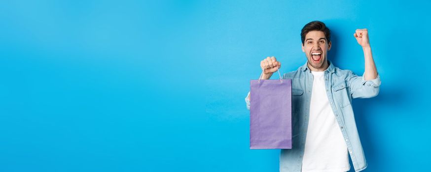 Concept of shopping, holidays and lifestyle. Cheerful young man celebrating, holding paper bag and making fist pump like winner, standing over blue background.