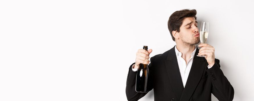 Close-up of handsome man in suit, kissing glass with champagne, getting drunk on a party, standing against white background.