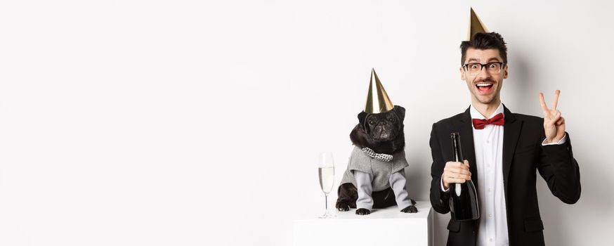 Small black dog wearing party hat and standing near happy man celebrating holiday, owner showing peace sign and holding champagne bottle, white background.