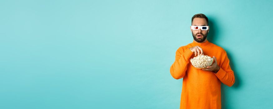 Image of intrigued man eating popcorn and watching movie in 3d, looking at tv screen, standing over blue background.