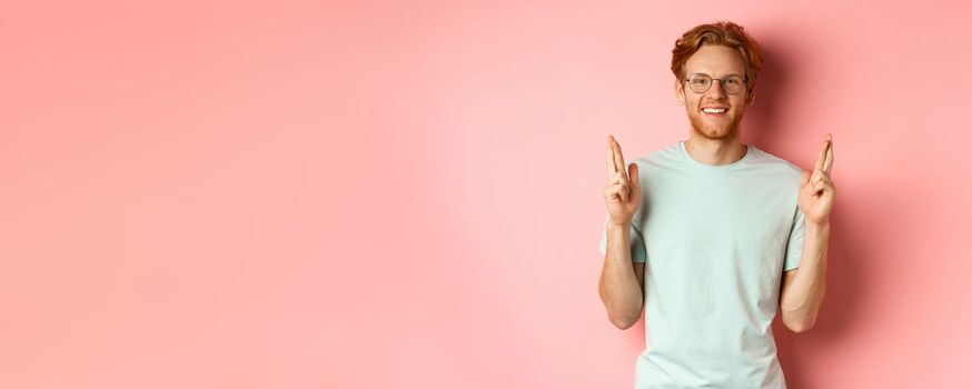 Attractive guy having faith in dreams, smiling hopeful and making wish with fingers crossed, feeling lucky, standing over pink background.