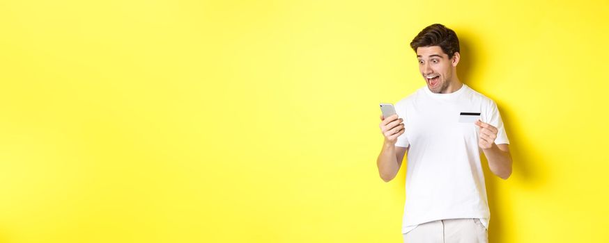 Man looking surprised at smartphone, shopping online, holding credit card, standing over yellow background. Copy space