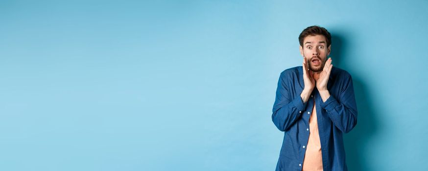 Shocked young man looking startled, screaming anxiously, standing on blue background. Copy space