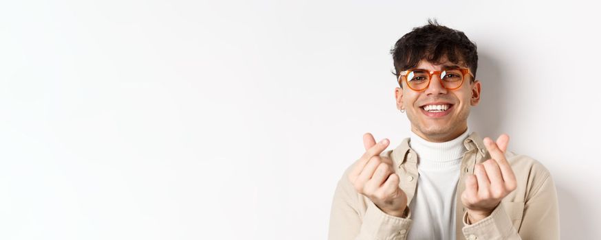 Cute young guy in glasses smiling and showing finger hearts, standing on white background.