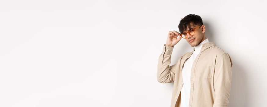 Handsome stylish guy saluting you, touching eyewear and smiling sassy, standing on white background.