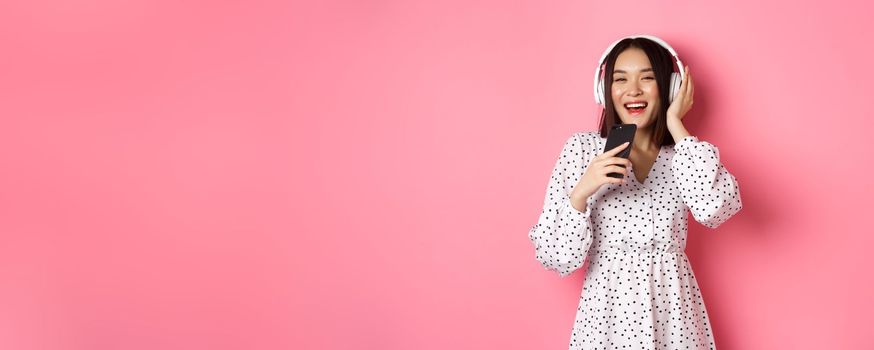 Beautiful smiling asian woman singing song in smartphone microphone, playing karaoke app and using headphones, standing over pink background.