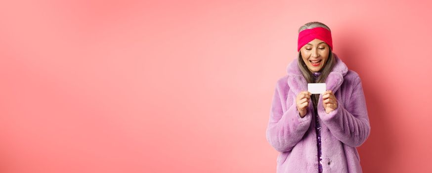 Shopping and fashion concept. Beautiful and stylish asian woman looking at plastic credit card, smiling pleased, standing over pink background.