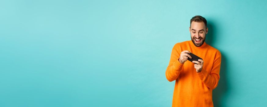 Excited adult man playing on mobile phone, looking amazed at smartphone screen, standing over turquoise background.