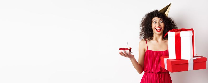 Cheerful girl in party hat and red dress, celebrating birthday, holding surprise gift and b-day cake, enjoying holiday, standing over white background.