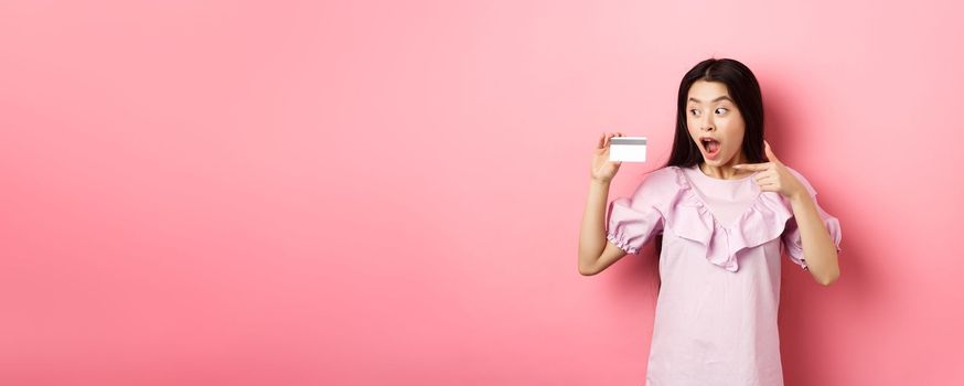 Excited asian woman pointing at plastic credit card, showing awesome advertisement, standing on pink background.
