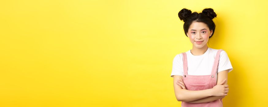 Beauty asian girl with glamour makeup, cross arms on chest and smiling tenderly at camera, standing on yellow background.