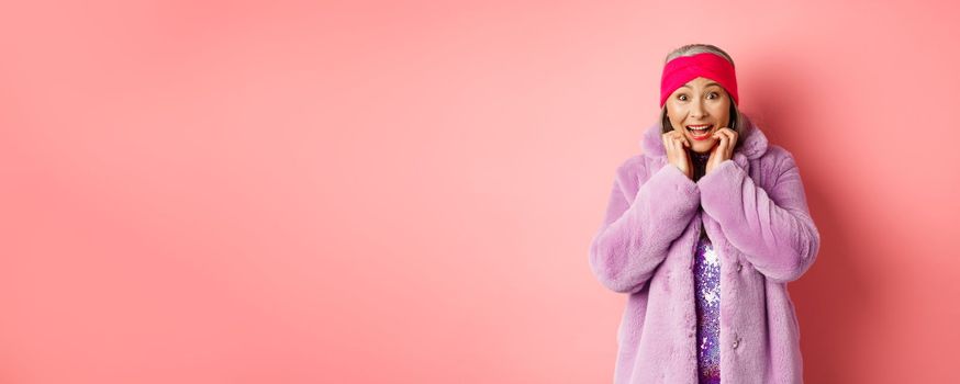 Excited and happy asian female senior model looking at camera with amazement, checking out promo offer, standing in trendy clothes over pink background.