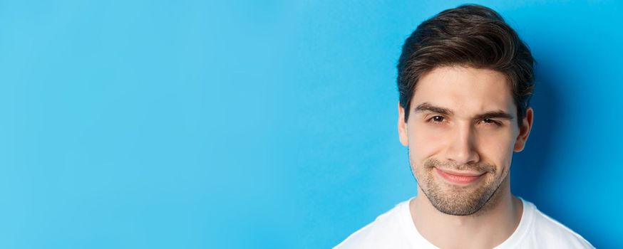 Headshot of thoughtful handsome man, looking intrigued and squinting, thinking about something, standing over blue background.