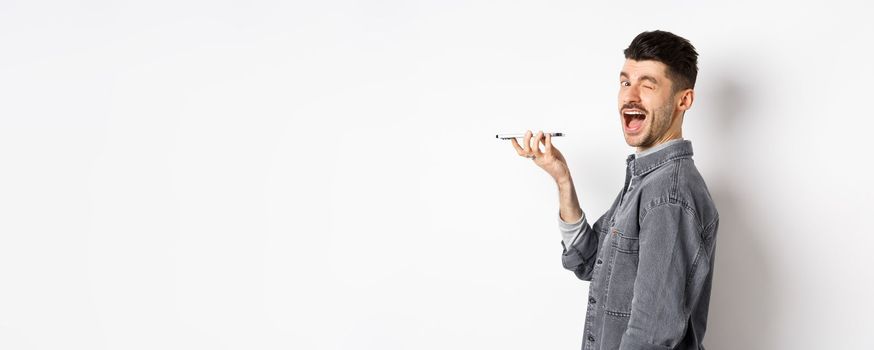 Happy guy winking at looking at camera while using voice translator app or talking on speakerphone, holding phone close to mouth, standing on white background.