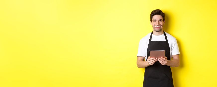 Waiter in black apron taking orders, holding digital tablet and smiling friendly, standing over yellow background.
