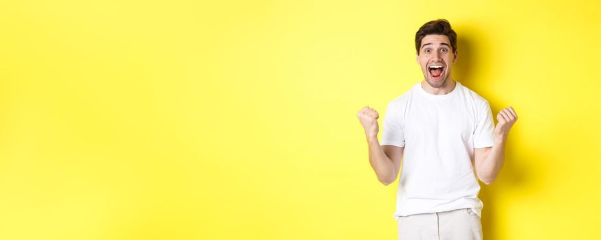 Excited and lucky man winning, clench fists and looking happy, triumphing and celebrating, standing over yellow background.
