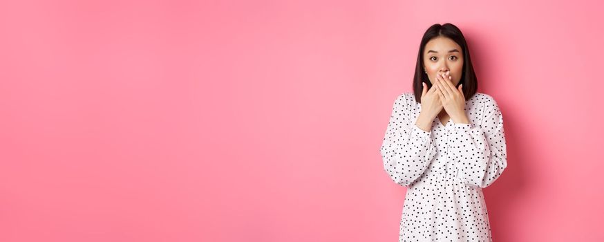 Image of surprised asian female model gasping, cover mouth and looking at camera, standing amazed against pink background.