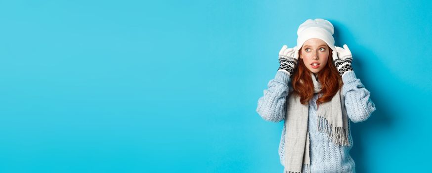 Winter and holidays concept. Cute redhead girl going outside, put on beanie and gloves, looking left, standing over blue background.