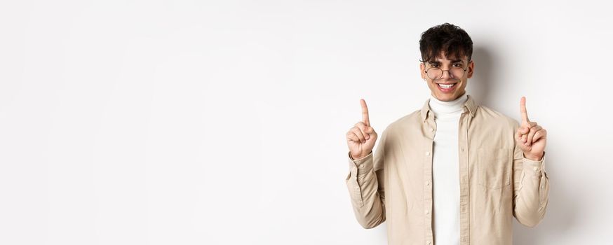 Portrait of handsome hipster guy in glasses showing advertisement and smiling, pointing fingers up at top offer, standing on white background.