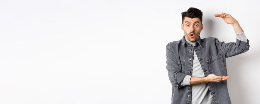 Cheerful guy smiling and showing big size, showing large thing and looking excited, standing against white background.