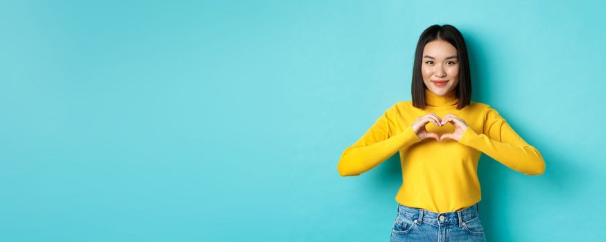 Attractive japanese girl in yellow sweater, showing heart gesture and say I love you, looking heartfelt at camera, standing over blue background.