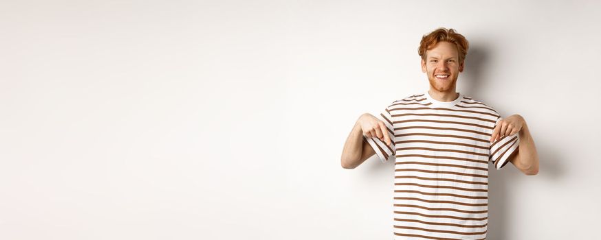 Smiling young man with messy red hair and beard, pointing fingers down, recommending shop, standing over white background.