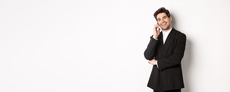 Image of handsome, successful businessman talking on phone, smiling pleased, standing in suit against white background.
