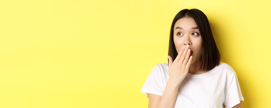 Close up of pretty asian girl looking shocked, gasping and covering mouth with hand, stare left at promotion, standing over yellow background.