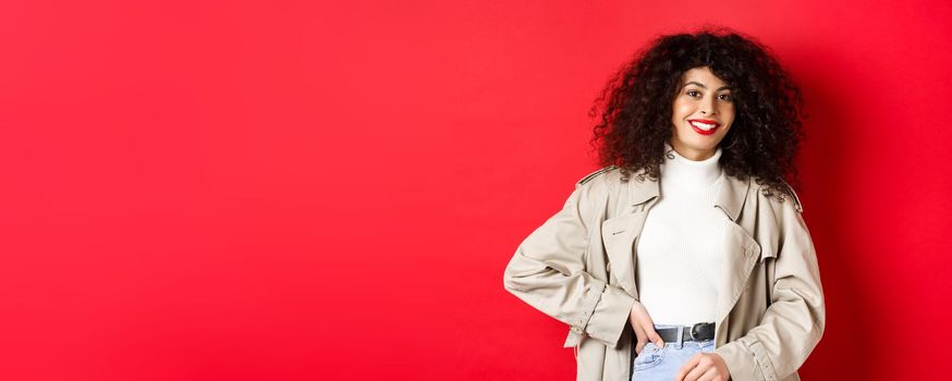 Modern woman with curly hair, wearing outdoor clothes, going for walk, standing against red background.