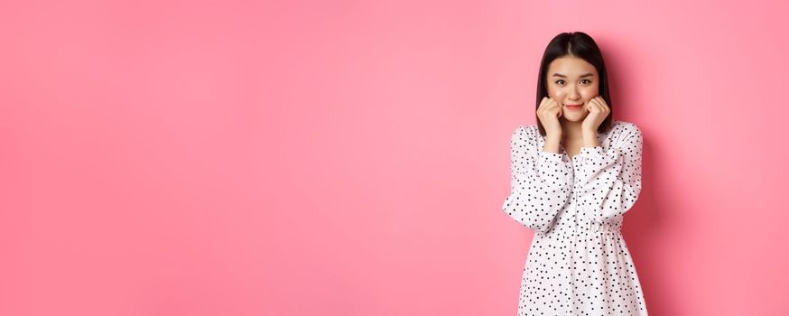 Cute and shy asian girl blushing, touching cheeks and looking at camera silly, standing against pink background.